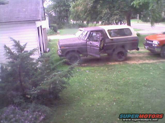 Purple Ford Bronco with open driver door, parked on grass next to a house. Another orange vehicle is visible to the right