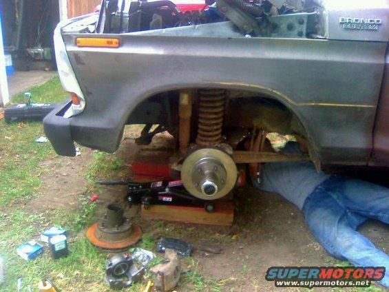 Gray Ford Bronco with front end disassembled, a person works underneath