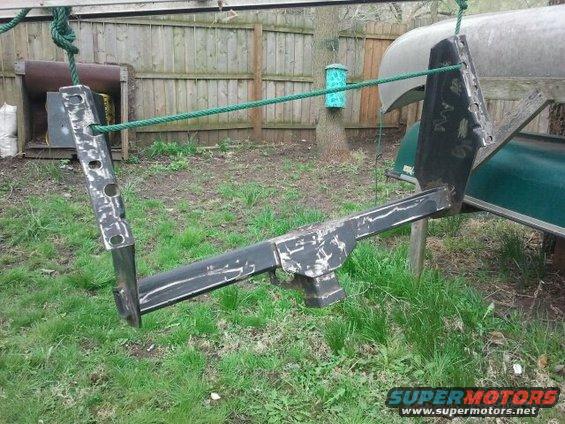 Black metal trailer hitch assembly hanging from ropes, with a canoe and fence in the background