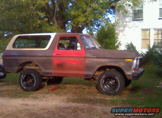 Side view of a rusty, two-tone Ford Bronco with red doors and a tan top. It has aftermarket wheels and appears to be lifted
