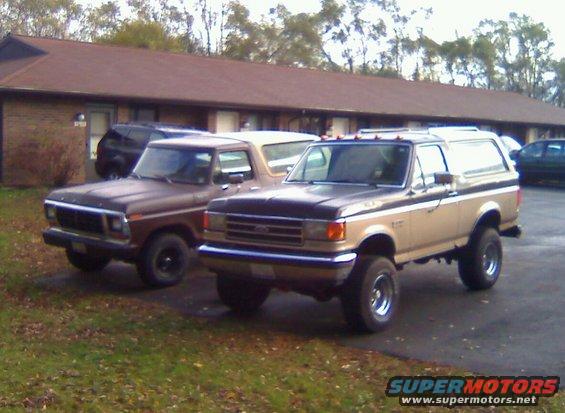 Two Ford Broncos parked side-by-side. The front Bronco is brown. The rear Bronco is tan with a black stripe and lifted suspension