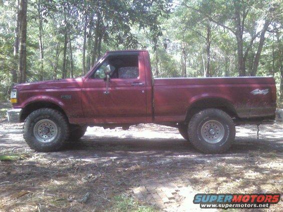 Maroon Ford F-150 pickup truck, side view, lifted suspension, off-road tires, parked in a wooded area