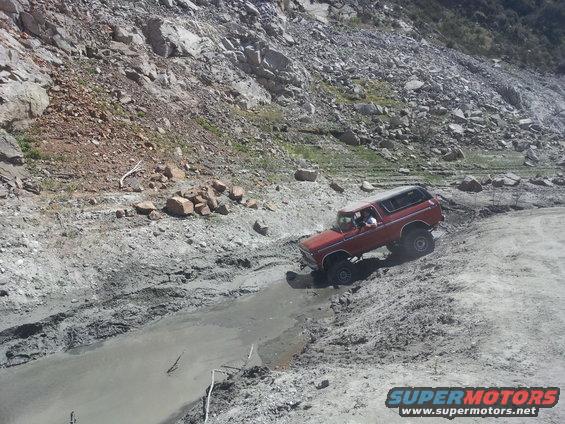 Red 1978 Ford Bronco driving off-road through a muddy ditch on a rocky slope