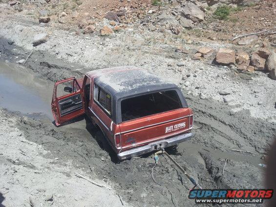 Red 1978 Ford Bronco stuck in mud with driver's door open, 'Alley Customs' decal on tailgate