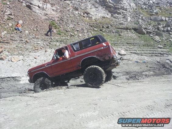 Red 1978 Ford Bronco stuck in mud, driver looking out window. Another person stands on hill behind it