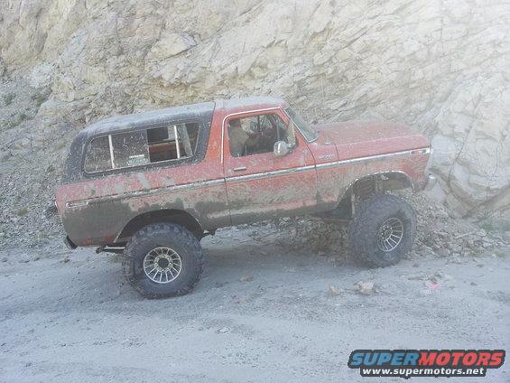 1978 Ford Bronco with lifted suspension and large tires driving on a dirt trail next to a rocky hill