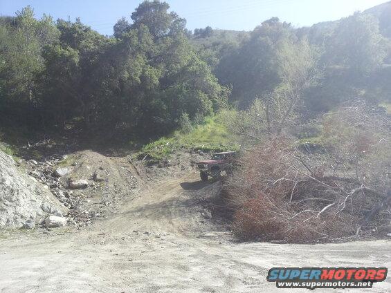 Red 1978 Ford Bronco driving up a dirt hill with trees and rocks