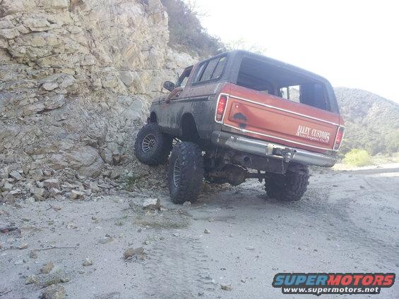Orange 1978 Ford Bronco with black top, lifted suspension, and large tires, driving on a dirt road next to a rocky incline