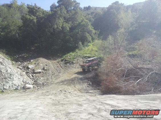 Red 1978 Ford Bronco driving up a dirt hill, front 3/4 view, some brush on the right