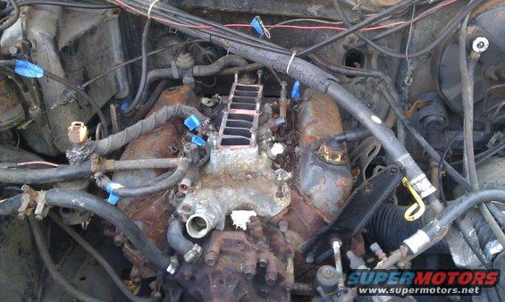 Engine bay with rusty intake manifold and hoses, some marked with blue tape