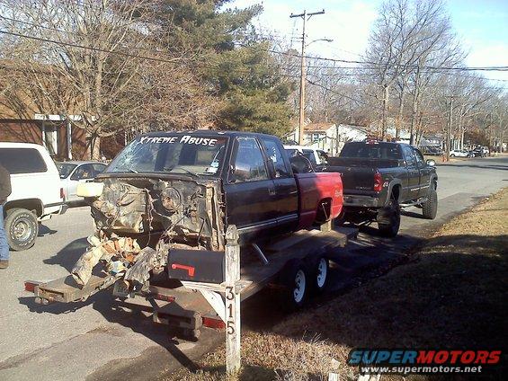 Pickup truck body on a trailer, front end is wrecked. Another lifted pickup is behind it. Text on windshield says 'XTREME ABUSE'