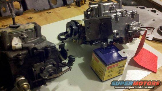 Two carburetors on a workbench, one appears older and darker, the other is shiny and silver. A blue and yellow box labeled 'Stant 13008' sits nearby