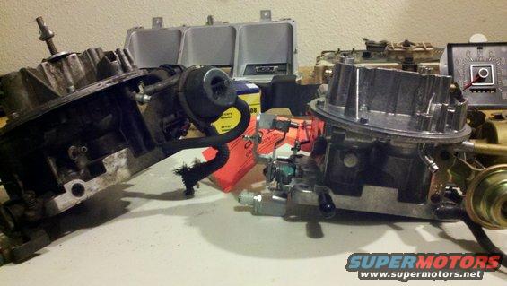 Two carburetors sit on a table with some other engine parts in the background