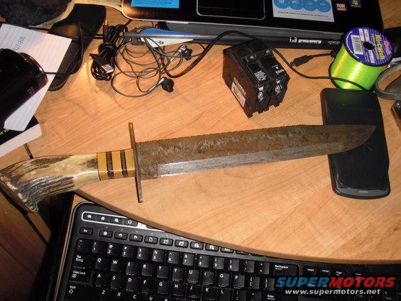 Large rusty knife with a serrated edge and an antler handle, lying on a wooden desk next to a keyboard and other objects