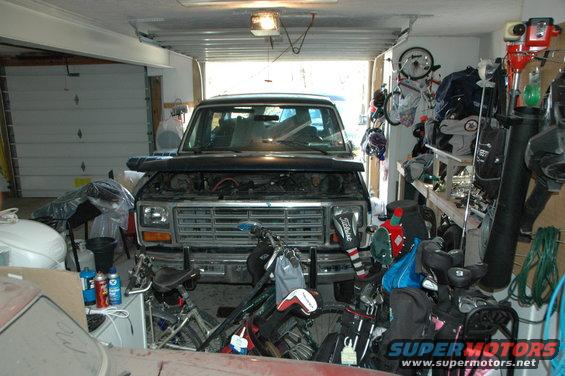 Front view of a dark-colored Ford Bronco with hood open, parked in a garage with bicycles and golf clubs