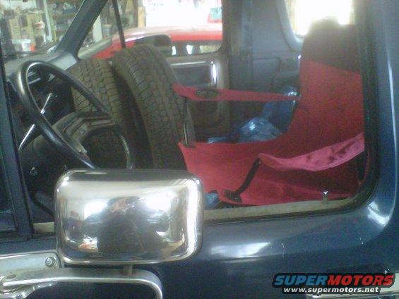 Interior view of a vehicle showing spare tires, a steering wheel, and red seats. Another vehicle is visible in the background