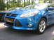 2012 Ford Focus - The front end of a blue Ford Focus is visible, parked on asphalt with trees in the background.