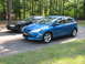 2012 Ford Focus - A blue 2012 Ford Focus hatchback is parked next to a black sedan in a driveway.