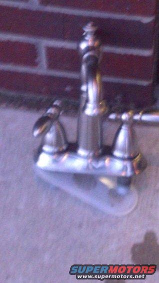 Chrome faucet with two handles and a central spout, sitting on a concrete surface with a brick wall behind it. Supermotors.net logo at the bottom