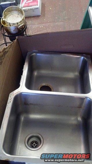 Double stainless steel sink in a cardboard box. A strainer basket and a box are visible behind it