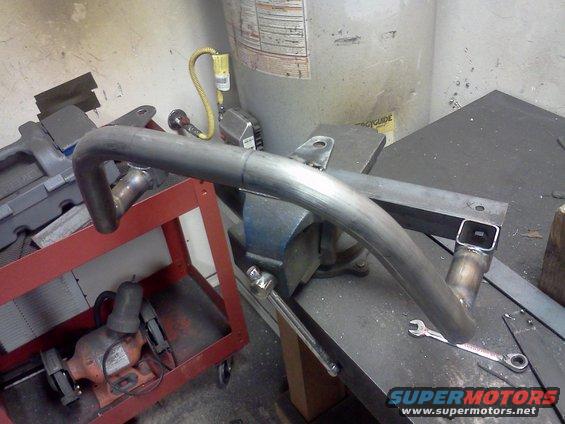Metal exhaust pipe section clamped in a vise on a workbench, with a grinder visible on a red cart