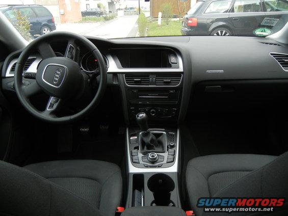 Interior view of an Audi with a steering wheel, dashboard, and center console visible. Another Audi is parked outside