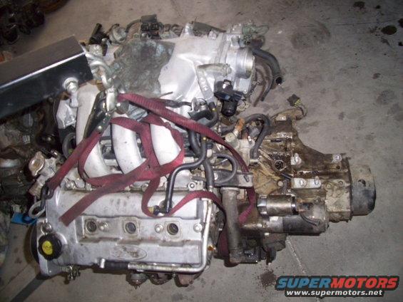 Engine with red straps, silver valve cover with Ford logo, and transmission attached