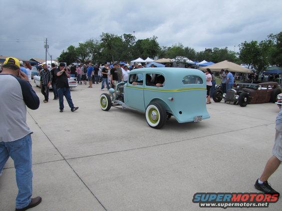 Light blue hot rod sedan with yellow trim, white wall tires, and exposed engine components, viewed from the rear