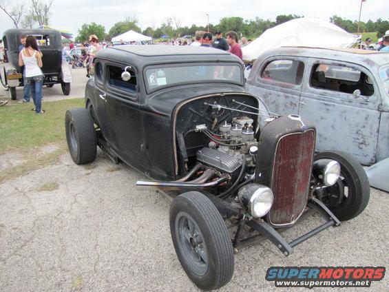 Black hot rod coupe with exposed engine and red grille, parked on gravel