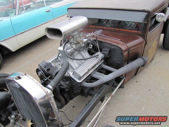 Hot rod engine with large scoop and exposed exhaust pipes. Another car is visible to the left