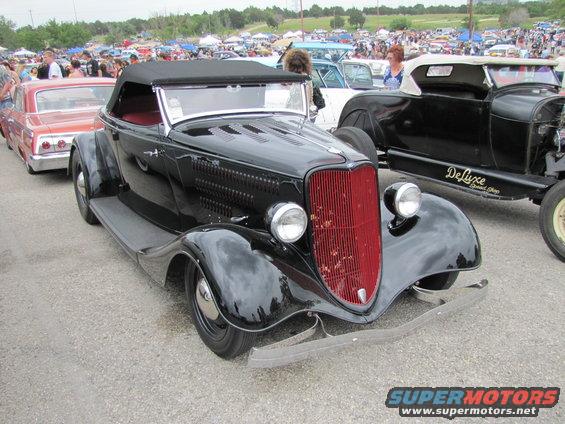 Black convertible hot rod with red grille and side exhaust, parked at an outdoor car show