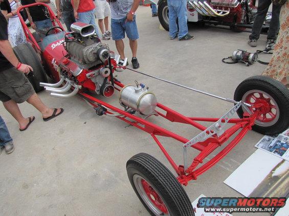 Red dragster chassis with a supercharged V8 engine, exposed exhaust pipes, and a fuel tank
