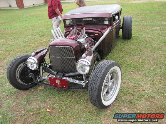 Maroon hot rod with exposed engine and chrome exhaust pipes, parked on grass