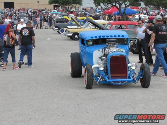 blue hot rod with exposed engine and red grille, parked at an outdoor car show with other vehicles and people