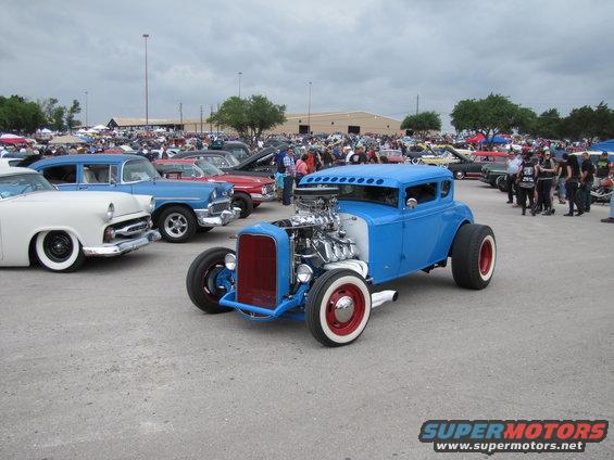 Blue hot rod with exposed engine, red grille, and white-wall tires parked at an outdoor car show