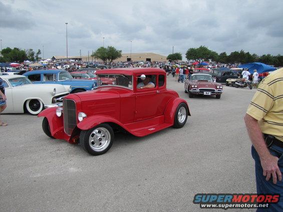 Red coupe hot rod with chrome wheels and a driver, angled from the front-left