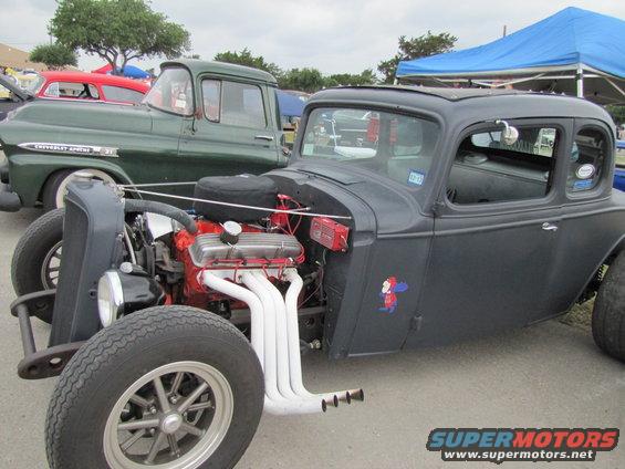 Hot rod coupe with V8 engine, custom exhaust, and a cartoon decal on the door. Green Chevrolet truck in background