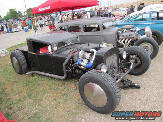 Custom hot rod with exposed V8 engine and turbocharger, parked next to a grey sedan and a blue car