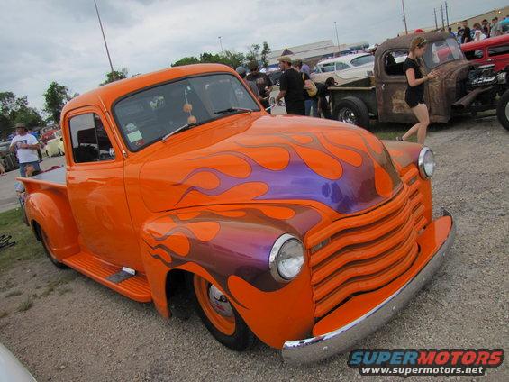 Orange pickup truck with purple and orange flames, chrome bumper, and horizontal grille. Another rusty truck is visible behind it