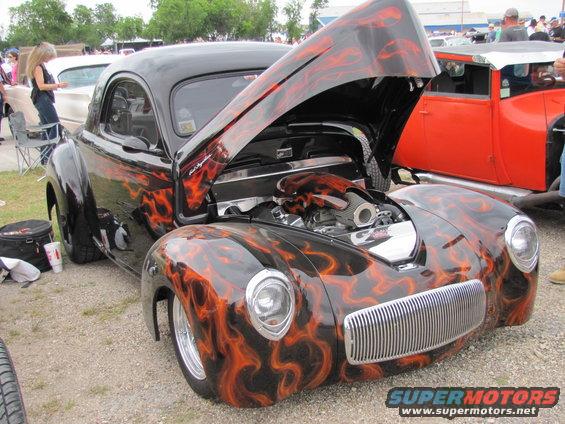 Black hot rod with orange flame graphics and hood open showing engine. Another orange car is parked next to it