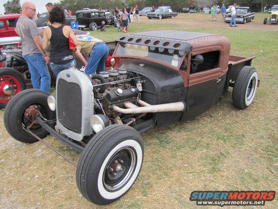 Custom hot rod pickup truck with exposed engine and whitewall tires. People are gathered around it