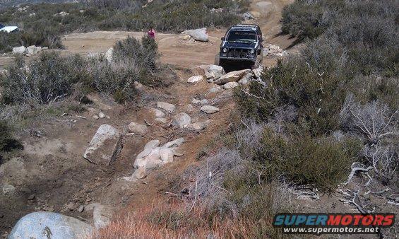 Black SUV with front end damage driving on a rocky off-road trail. A person stands in the background