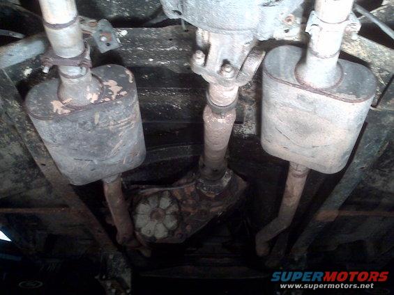 Underside view of a vehicle's dual exhaust system, showing mufflers and driveshaft