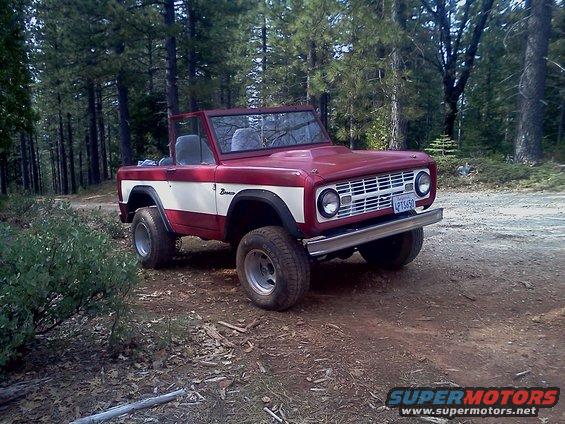 1968 Ford Bronco parked on a dirt road in a forest, red with white stripe