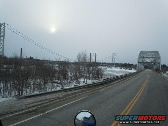 Truck cab mirror reflecting blue truck, driving on a road towards a bridge in winter