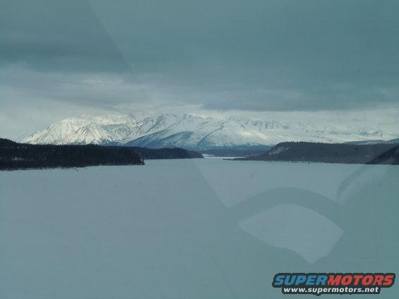 Snowy mountains overlook a frozen lake under a cloudy sky. Watermark reads SUPERMOTORS