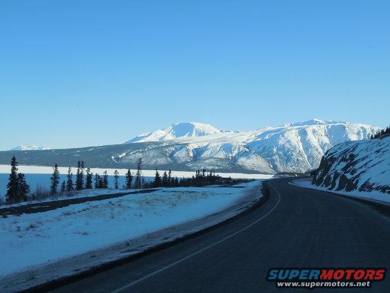 road curves through snowy mountains with a frozen lake on the left