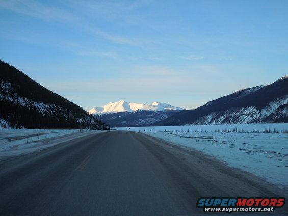 Winter road leading towards snow-capped mountains under a clear blue sky. Snow covers the ground and trees