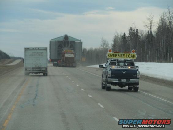 Pickup truck with 'OVERSIZE LOAD' sign, escorting two semi-trucks on a highway