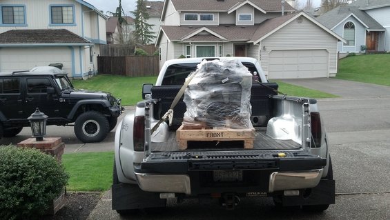 Black Jeep Wrangler parked on street, with a silver pickup truck carrying an engine on a pallet in its bed